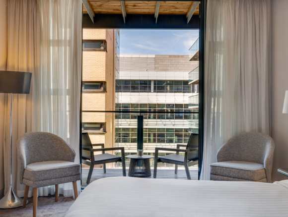 Bright hotel room with king bed, two armchairs and balcony seating overlooking modern city buildings, sheer curtains and bedside lamps
