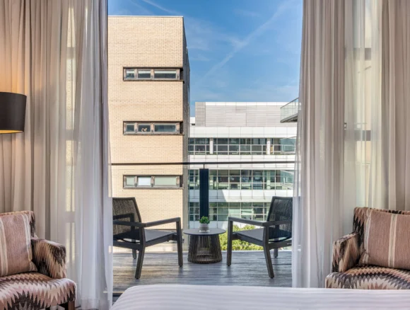 City-view hotel room with sheer curtains, balcony seating, small table and modern lamp — ideal for business or leisure stays