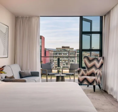 Light-filled hotel suite with king bed, sofa, patterned armchair and balcony seating overlooking city view and modern amenities