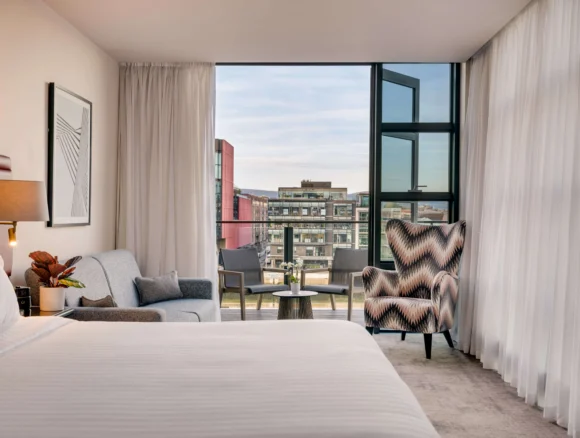 Light-filled hotel suite with king bed, sofa, patterned armchair and balcony seating overlooking city view and modern amenities