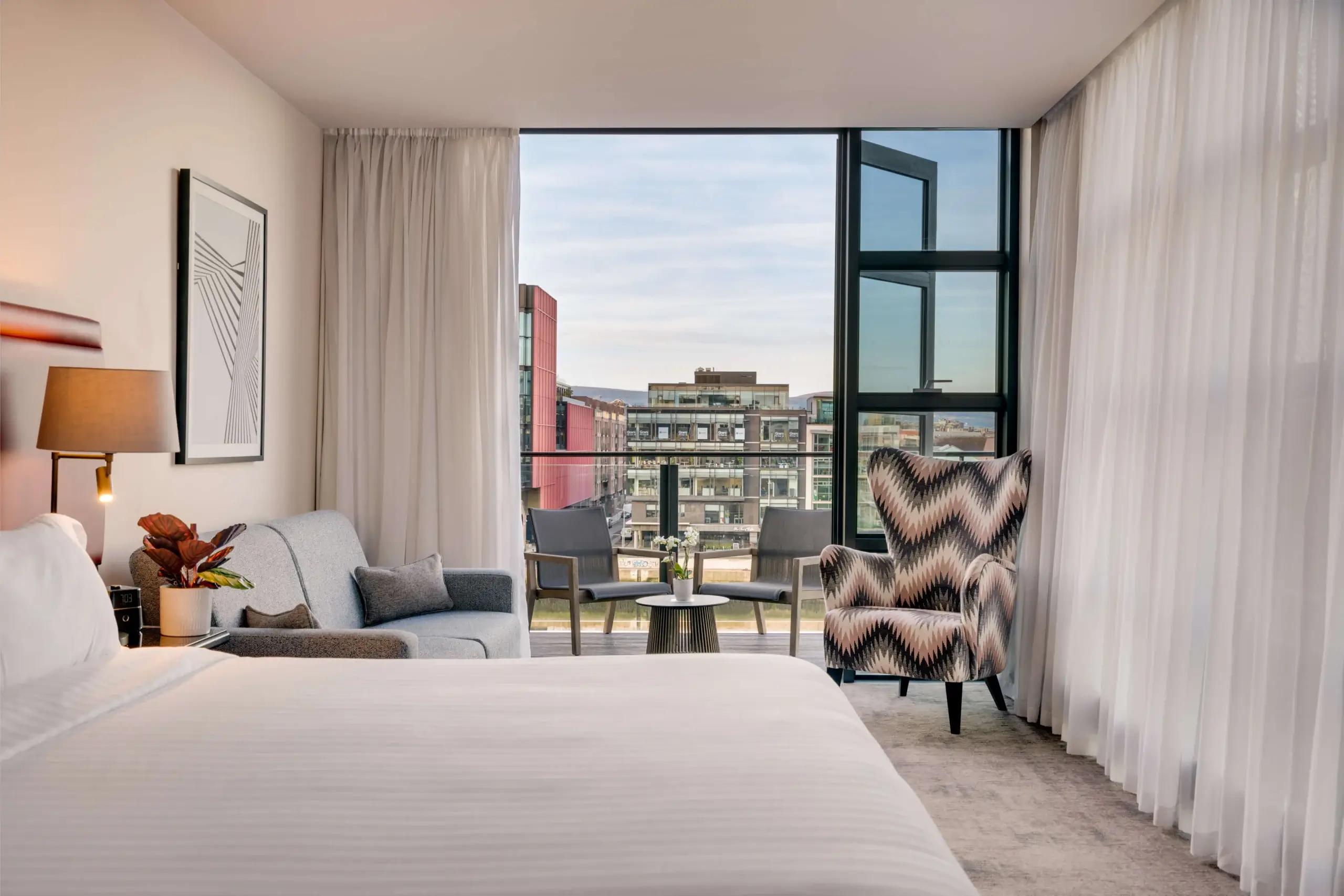 Light-filled hotel suite with king bed, sofa, patterned armchair and balcony seating overlooking city view and modern amenities