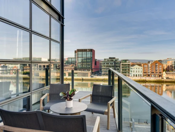 Riverside balcony with table, chairs and plant, glass railing overlooking modern waterfront buildings and river views, welcoming hotel amenity