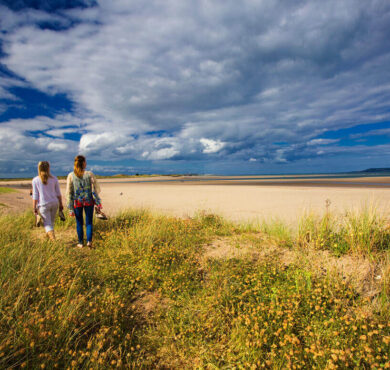 Guests enjoying scenic beach walk with lush dunes under a vibrant sky near luxurious seaside hotel.