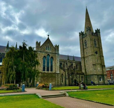 Historic stone cathedral with tall spire, lush green lawns, and seated visitors enjoying the serene park setting.