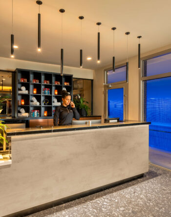 Welcoming hotel reception with indoor pool view and modern décor, featuring stylish lighting and lush plants.