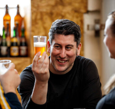 Guests enjoying craft beer tasting experience in cosy hotel bar with rustic decor, bottled selection in background.