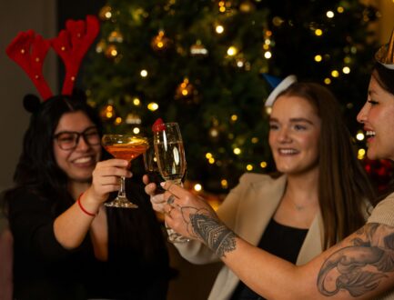 Guests celebrating with festive drinks and party hats in front of a sparkling Christmas tree at a cosy hotel.