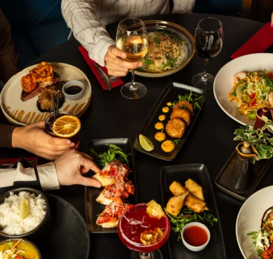 Overhead view of shared hotel dining: seafood salad, steak, tapas and wine glasses on a black table, relaxed restaurant vibe