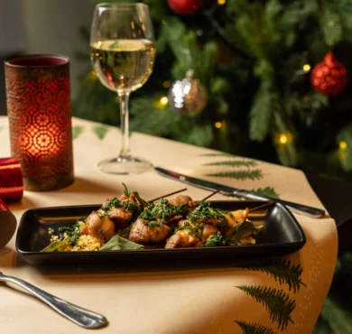 Festive hotel dinner table with skewered chicken, wine glass, lit candle and decorated Christmas tree in background