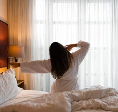 Guest in a white robe stretching on a king bed by floor-to-ceiling sheer curtains, warm bedside lamp and plush pillows visible — relaxing city-stay room