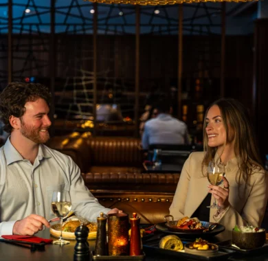 Couple enjoying evening hotel dining with wine, plated meals and cosy leather banquette in a dimly lit bar-restaurant